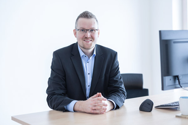 Jörn Hillmer sitzt an einem Schreibtisch und lächelt in die Kamera.