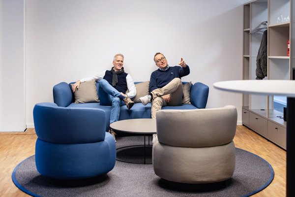 Marc Ellinghaus und Christian Buß sitzen auf einem blauen Sofa. Marc zeigt in die Ferne, Christians Blick folgt.