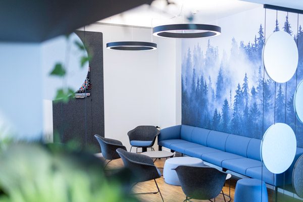 Aufenthaltsraum der HEB Steuerberatung mit einem langen, blauen Sofa, grauen Sitzmöbeln und einer Fototapete mit einem blauen Wald